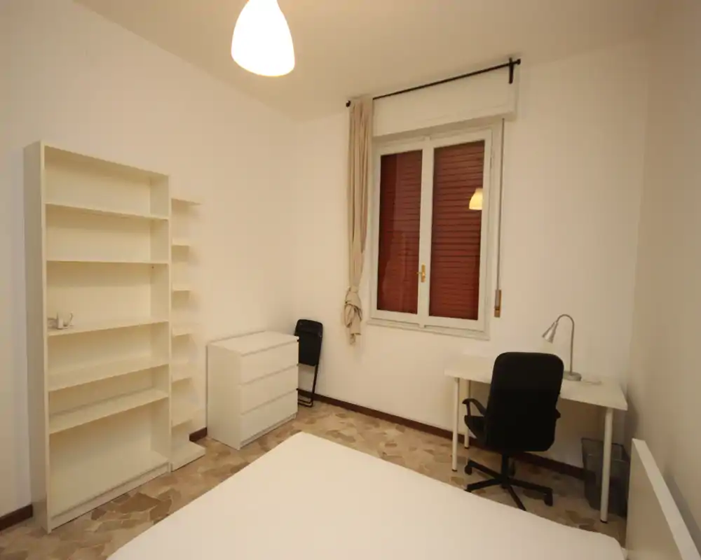 Camera da letto luminosa e arredata in modo semplice con letto in primo piano, scaffali bianchi e una scrivania vicino alla finestra — pulita e funzionale sia per soggiorni brevi che lunghi.