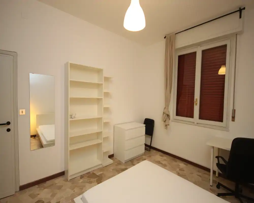 Camera da letto semplice e ben illuminata con letto matrimoniale visibile, scaffalatura, comò e scrivania. Arredo neutro e pavimento in piastrelle, ideale per affitti brevi.