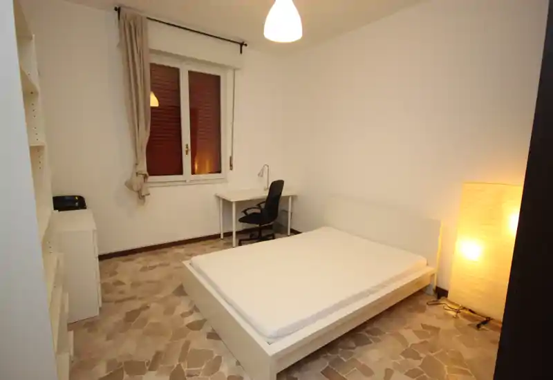 Camera da letto minimal e moderna con letto matrimoniale bianco, lampada da terra accanto al letto e una piccola scrivania vicino alla finestra. Toni neutri e luce calda creano un ambiente semplice e confortevole, ideale per ospiti o studenti.