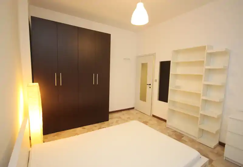 Chambre lumineuse et sobrement meublée avec lit double au premier plan, grand placard foncé et étagères blanches. Lampe sur pied chaude et plafonnier apportent une atmosphère accueillante ; sol carrelé et murs blancs pour un rendu propre et minimaliste.