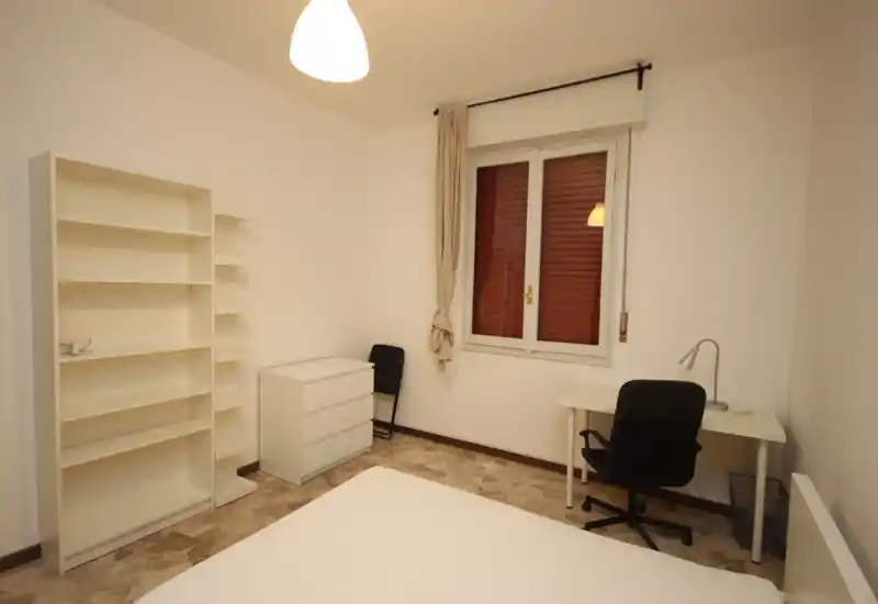 Camera da letto luminosa e arredata in modo semplice con letto in primo piano, scaffali bianchi e una scrivania vicino alla finestra — pulita e funzionale sia per soggiorni brevi che lunghi.