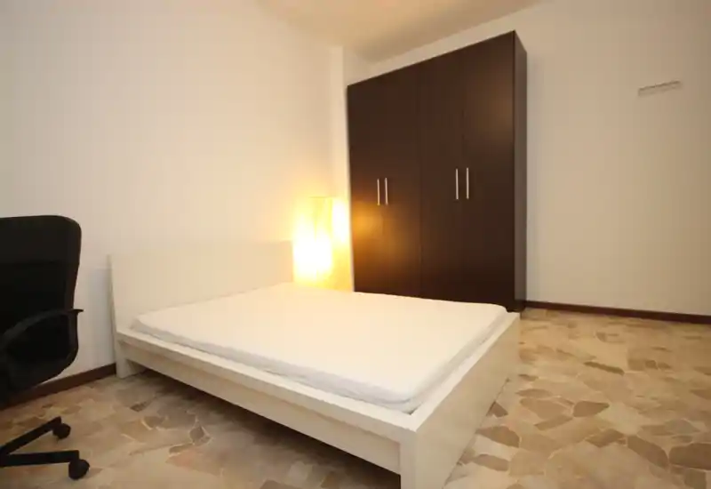 Camera da letto minimale e ordinata con letto bianco basso, armadio scuro e una lampada da terra che crea un'illuminazione calda.