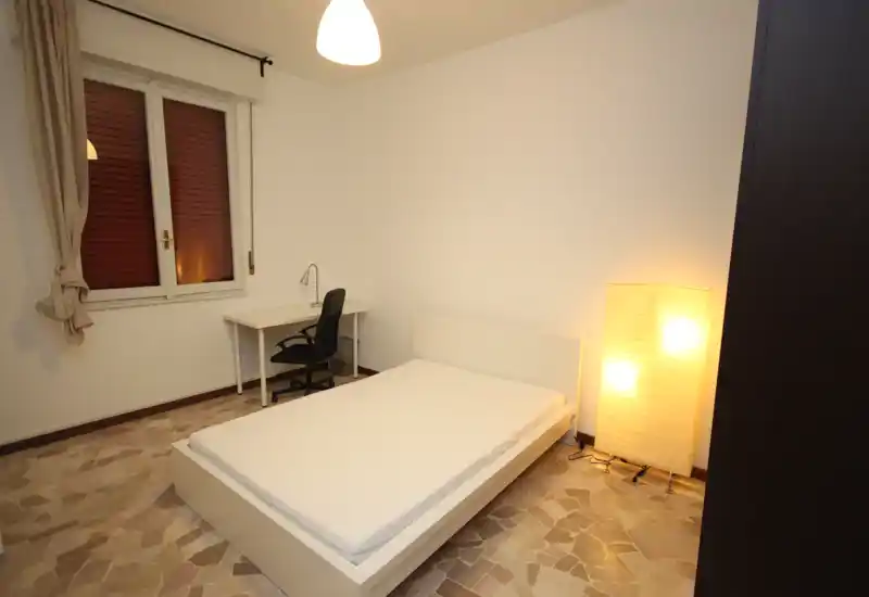 Camera da letto minimale con letto matrimoniale su piattaforma bianca, lampada da terra accanto e una piccola scrivania sotto la finestra — arredo semplice e luce calda.