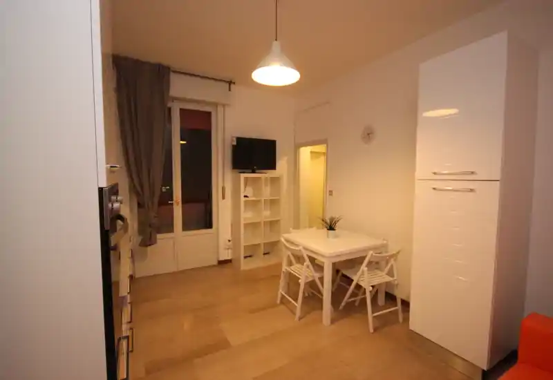 Compact open-plan dining area with a small white table and four folding chairs, a pendant light and shelving — ideal for casual meals in a modern rental.