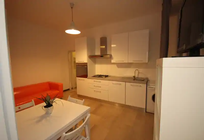 Modern open kitchen with white cabinets, a gas hob, built-in oven and sink; small dining table and orange sofa create a cozy studio layout.