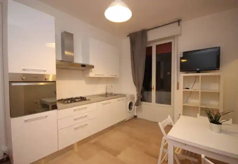 Modern open kitchen with white cabinetry, built-in oven, gas cooktop and integrated sink; small dining table and washing machine by the balcony door create a functional, bright space.