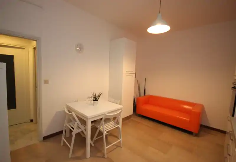 Compact living area with a bright orange two-seater sofa, small white dining table and chairs, and simple modern lighting — ideal for a studio or small apartment.