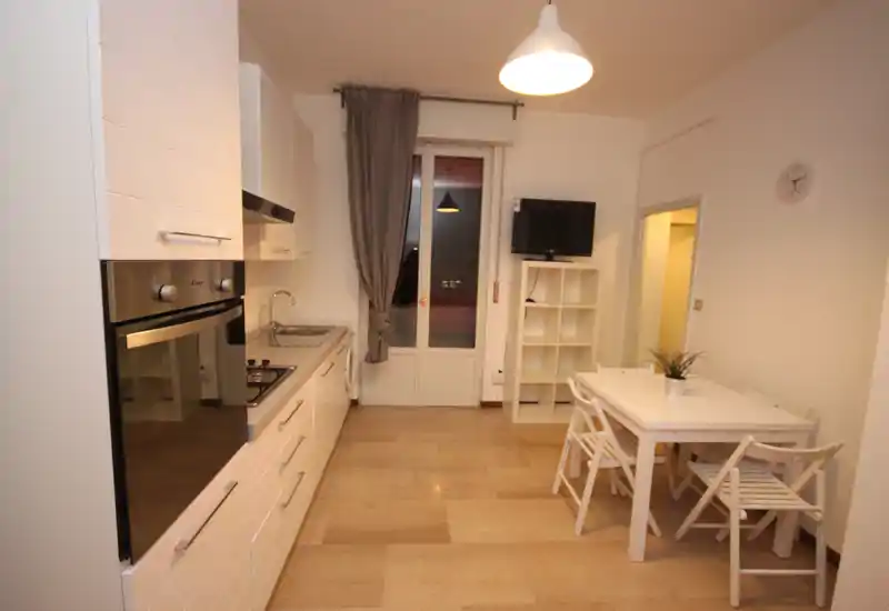 Modern open kitchen with built-in oven, stovetop and a small dining table. Neutral tones, light wood floor and a curtained door to a balcony create a bright, functional space.