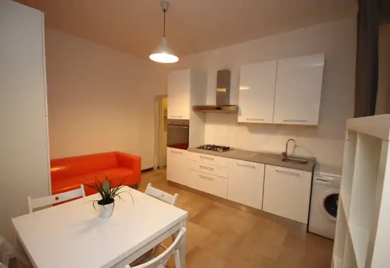 Compact modern kitchenette with white cabinets, gas hob, sink and a small dining table; an orange sofa adds a colorful accent.