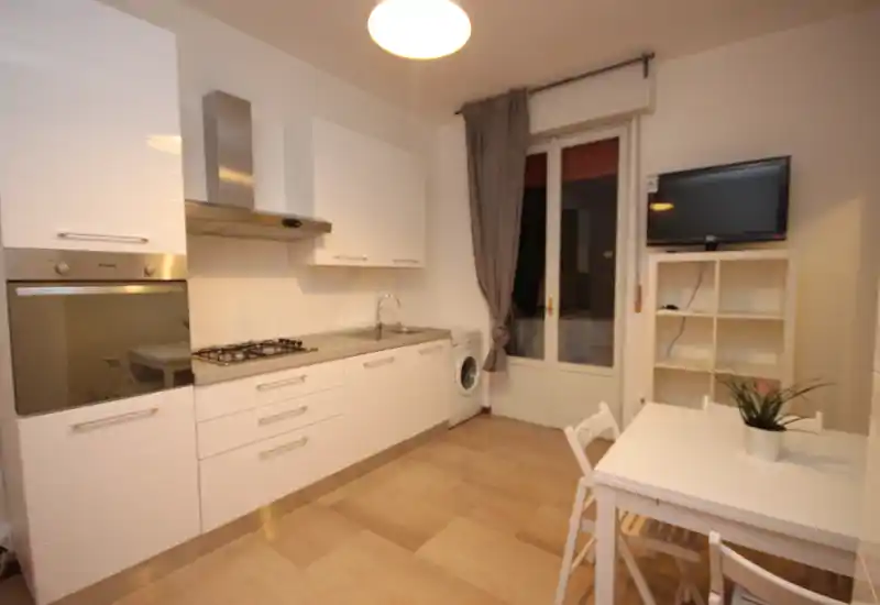 Modern, compact kitchen with white cabinetry, built-in oven, gas hob, sink and a small dining table. Bright, tidy space with a washing machine and balcony/door access, ideal for a functional rental kitchen.