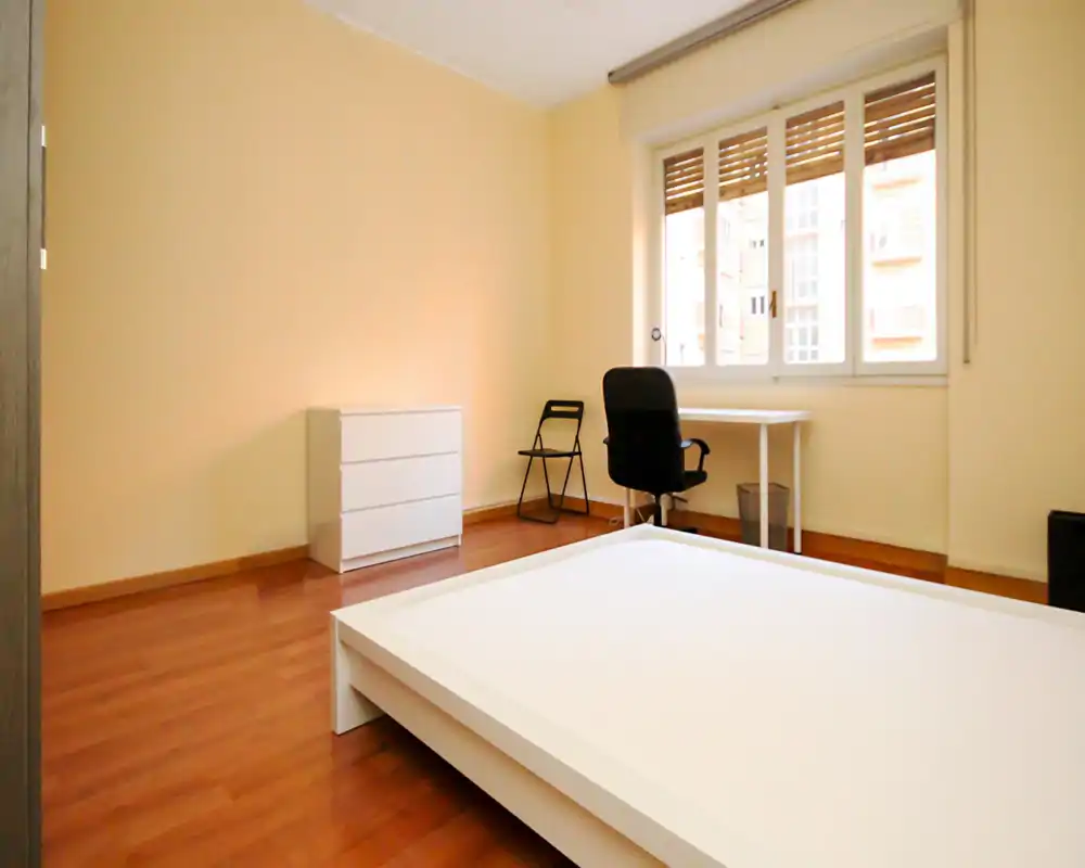 Bright, minimal bedroom with a low white platform bed, wooden floors and a desk by a large window providing abundant natural light.