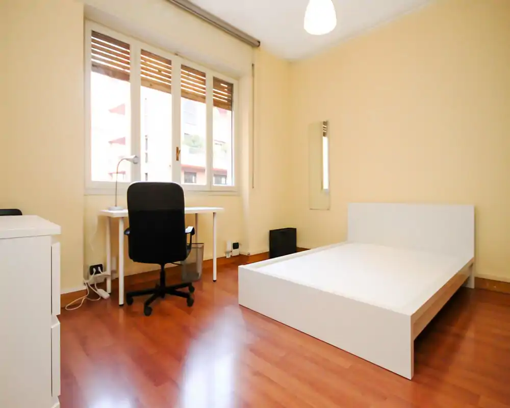 Bright, minimal bedroom with a white platform bed, wooden floors and a desk by the large window—ideal for a student or professional looking for a clean, functional room.