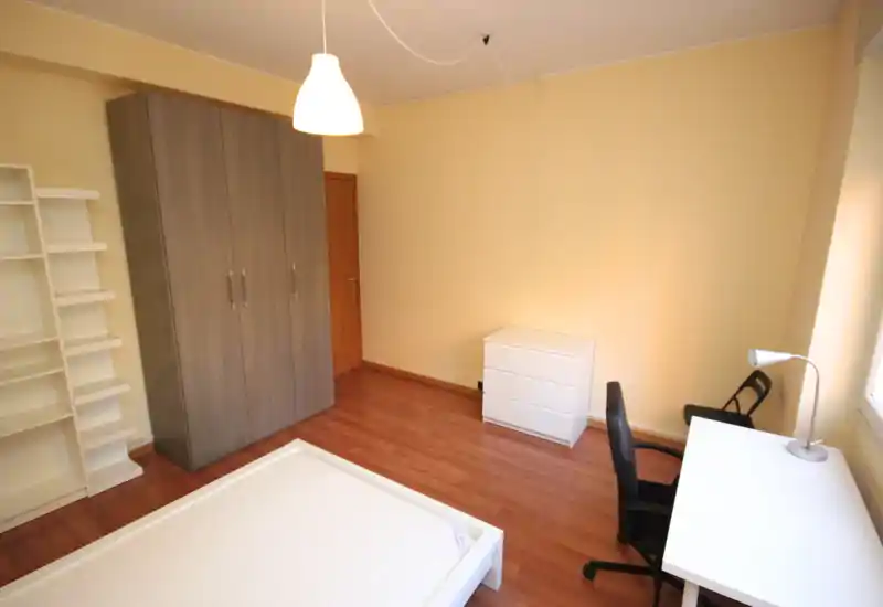 Simple, well-lit bedroom with a visible double bed frame, wardrobe, shelving and a work desk by the window — ideal for student or single occupancy.