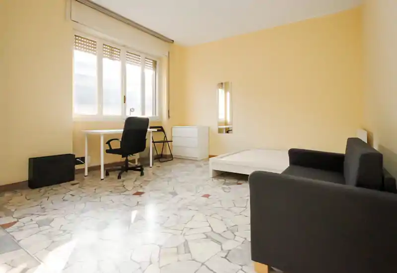 Camera da letto luminosa e semplice con rete letto bianca bassa, scrivania vicino a una finestra ampia e un piccolo divano — ambiente arioso con pavimento in piastrelle e arredi minimal.