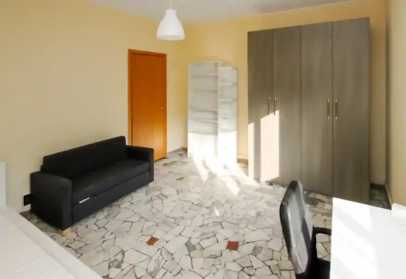 Camera da letto luminosa e arredata in modo essenziale con materasso visibile in basso a sinistra, divano nero, armadio e scaffalatura; la stanza è illuminata naturalmente e ha pavimento in pietra a mosaico.