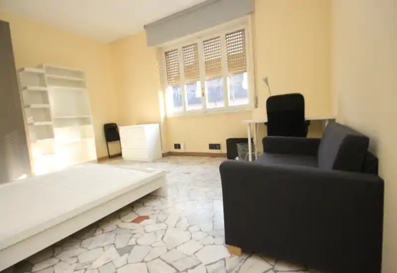 Dormitorio luminoso y amueblado de forma sencilla con estructura de cama baja visible, pequeño sofá, escritorio y estantería; gran ventana con luz natural intensa.