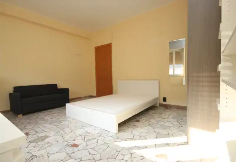 Dormitorio luminoso y minimalista con cama plataforma blanca y sofá oscuro de dos plazas; suelo efecto mármol y luz natural desde la derecha.