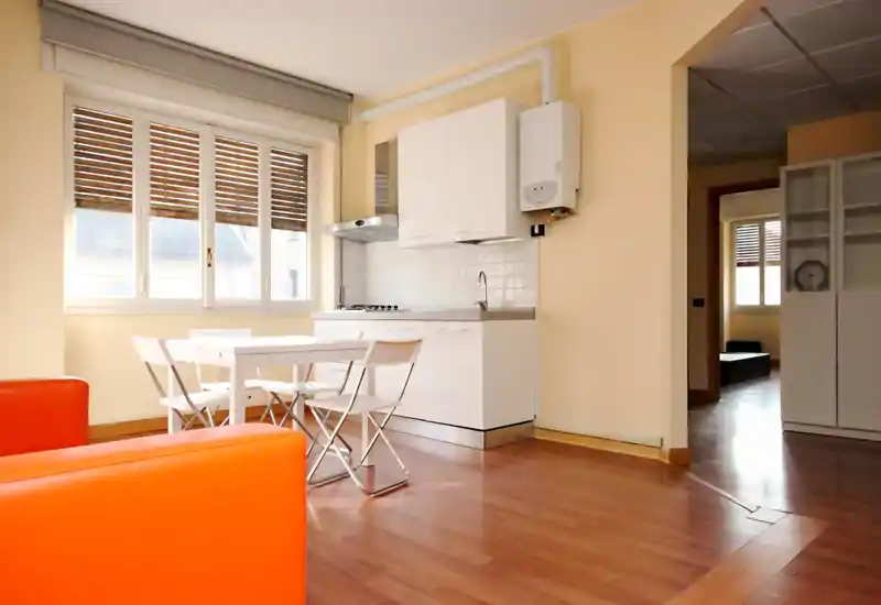 Zona giorno luminosa e moderna con angolo cottura bianco e tavolo da pranzo; pavimenti in legno caldo e un divano arancione che aggiungono colore e accoglienza.