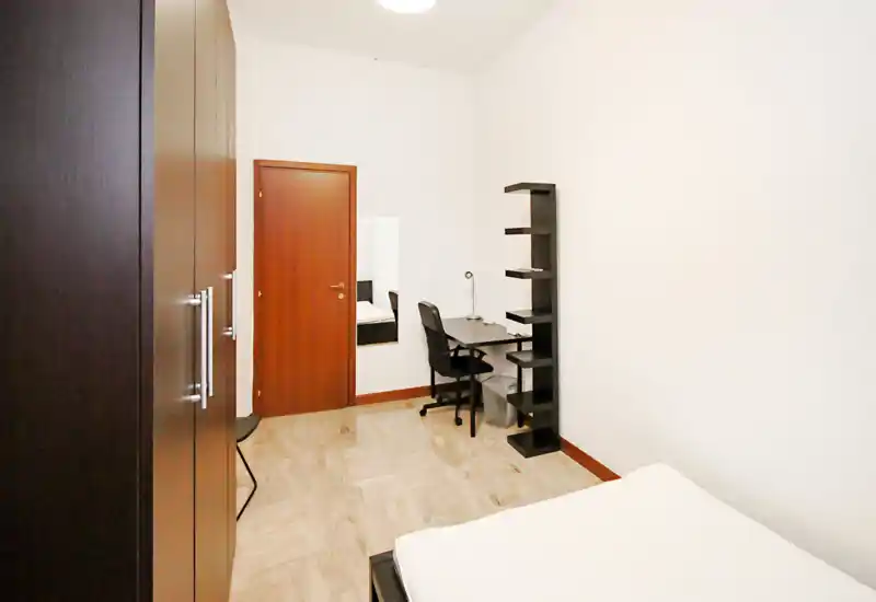 Bright, minimally furnished bedroom with a visible bed in the foreground, a wardrobe to the left and a small desk and shelving near the door — clean, modern lines ideal for a single occupant.