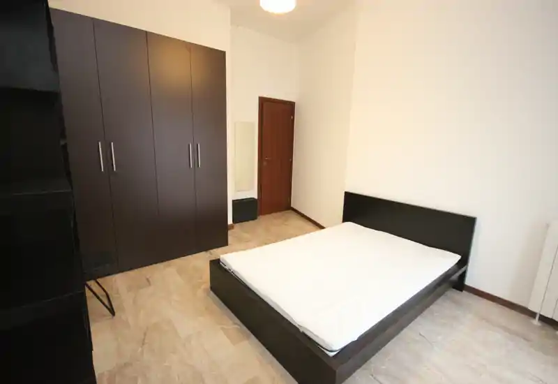 Dormitorio espacioso con cama de estructura oscura y gran armario; suelos neutros y paredes claras crean un espacio limpio y acogedor.