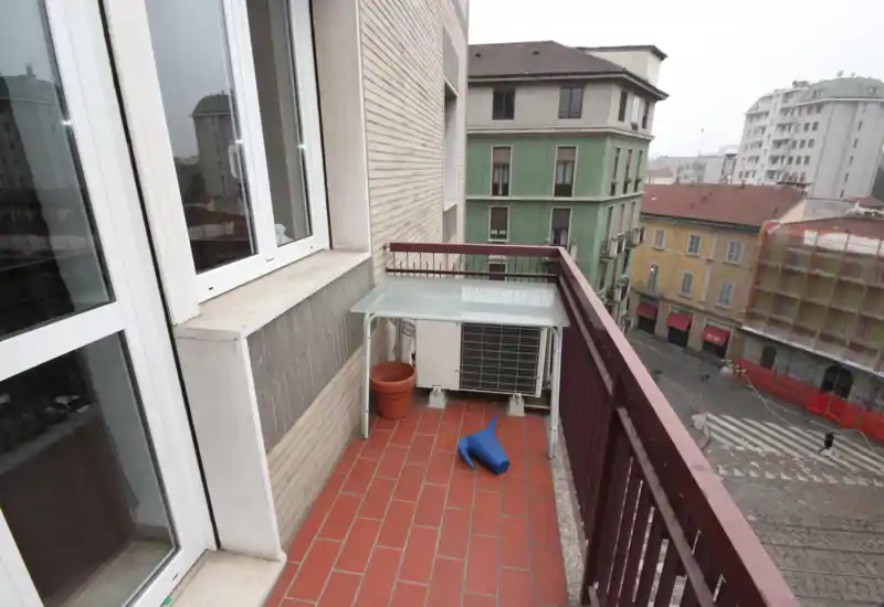 Small city balcony with terracotta floor tiles, metal railing and an external AC unit. The view overlooks neighboring buildings and a street, suitable for a compact outdoor seating area.