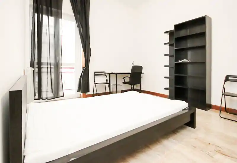 Dormitorio luminoso y minimalista con cama grande, escritorio y estantería. La luz natural atraviesa las cortinas oscuras translúcidas, aportando una sensación moderna y limpia.