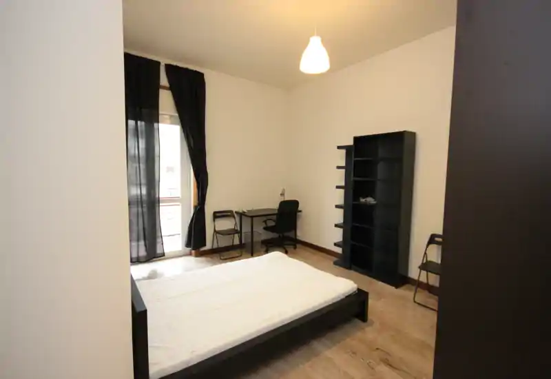 Minimal, well-lit bedroom with a low double bed, desk and shelving; neutral décor and natural light from a balcony window.