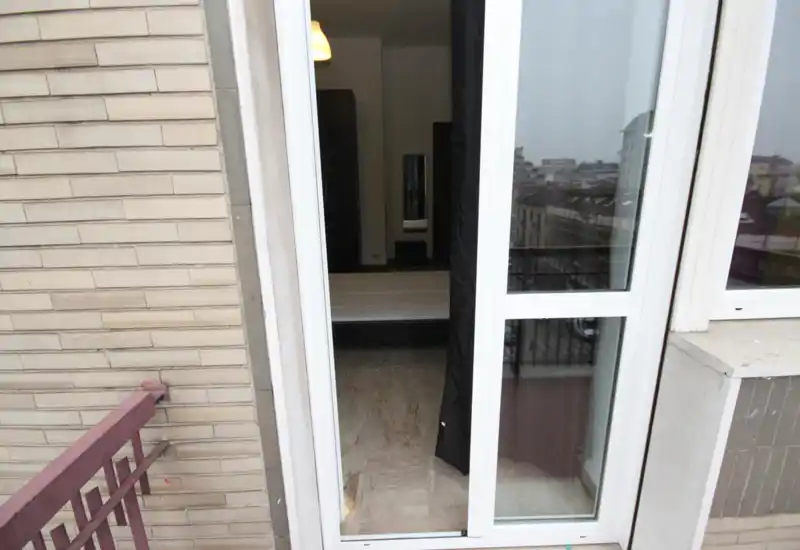 View from a balcony into a bedroom through sliding glass doors; a low-profile bed is visible inside with neutral flooring and minimal decor.