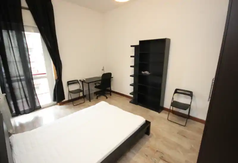 Simple, bright bedroom with a double bed, desk and shelving; balcony access with dark curtains provides natural light.