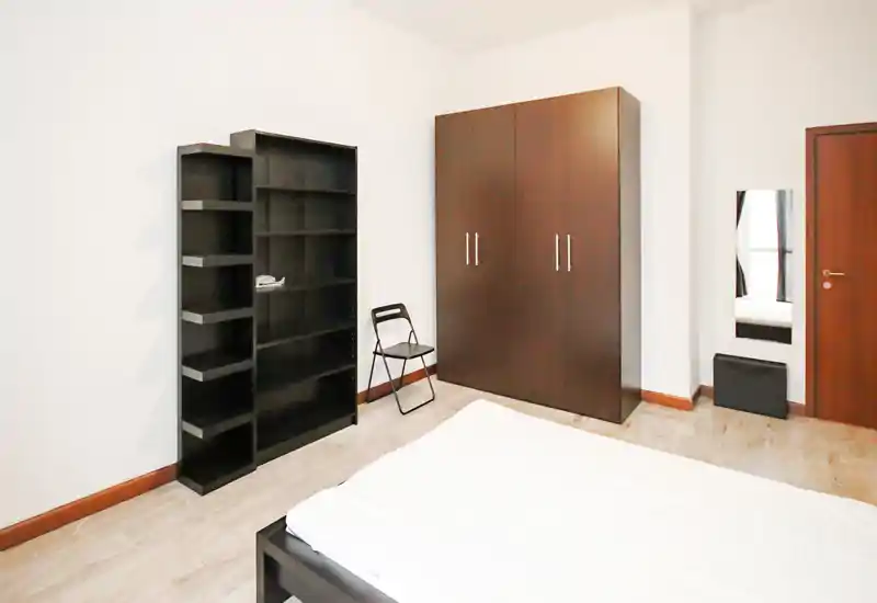Bright, minimal bedroom with visible bed, large wardrobe and shelving; simple modern furnishings and neutral tones create a calm, functional space.