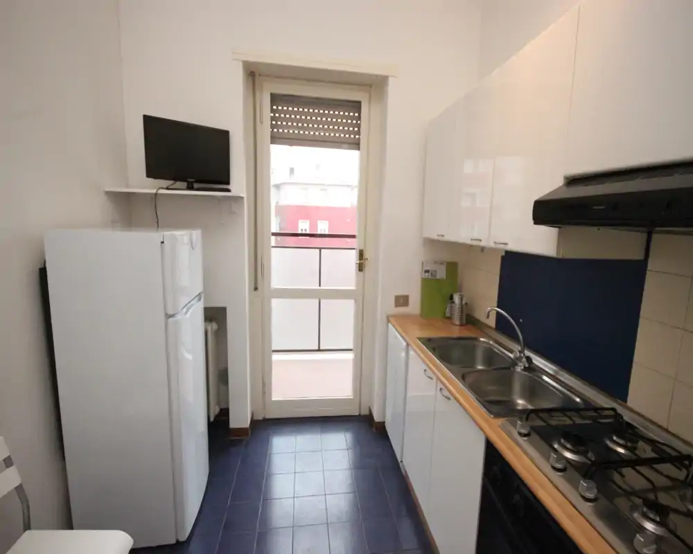 Cocina compacta y funcional con placa de gas, fregadero doble y encimera de madera. Luz natural a través de la puerta que da a un pequeño balcón.