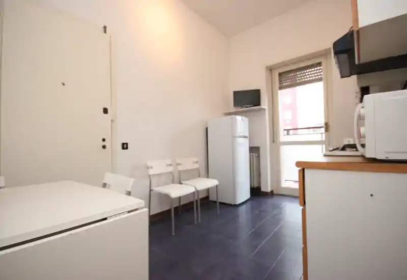 Compact kitchen with white cabinets and appliances, a small fridge and dining surface beside a balcony door providing natural light; minimal, functional layout ideal for short-stay rentals.