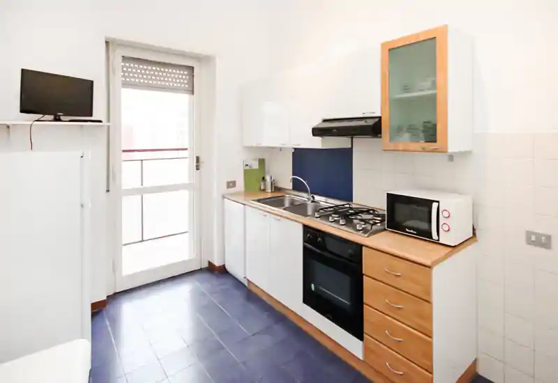 Compact, bright kitchen with white cabinets, wooden countertops and integrated gas hob. A door to a balcony provides natural light and the layout includes a sink, oven and microwave — practical and well maintained.