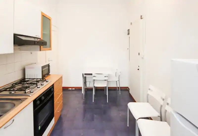 Compact kitchen with gas hob, oven and wall cabinets; small foldable dining table and chairs. Bright neutral walls and simple, functional layout ideal for a studio or small apartment.