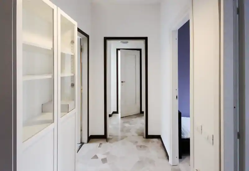 Pasillo/entrada luminoso y limpio con suelo de baldosas y varias puertas; estantería empotrada a la izquierda y una puerta hacia un dormitorio a la derecha.