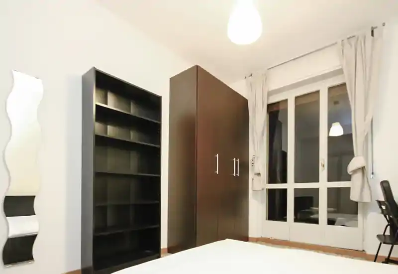Dormitorio moderno y minimalista con la cama en primer plano, un armario alto oscuro y estanterías, y una gran ventana/puerta con cortinas claras—líneas limpias y tonos neutros.