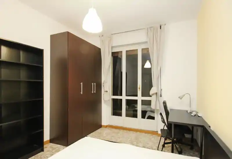 Dormitorio sencillo y bien iluminado con armario, estantería y escritorio junto a la ventana — práctico y despejado, ideal para estudiante o una sola persona.