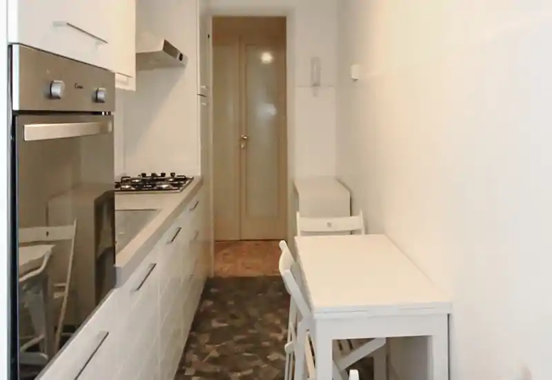 Cocina tipo galería estrecha con muebles blancos modernos, horno de acero inoxidable y una pequeña mesa — diseño compacto y funcional ideal para estudios o pisos pequeños.