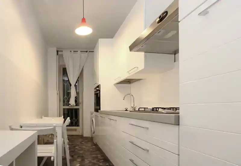 Cocina moderna y estrecha con muebles blancos, fregadero de acero, placa de gas y pequeña mesa; luminosa, limpia y bien distribuida.