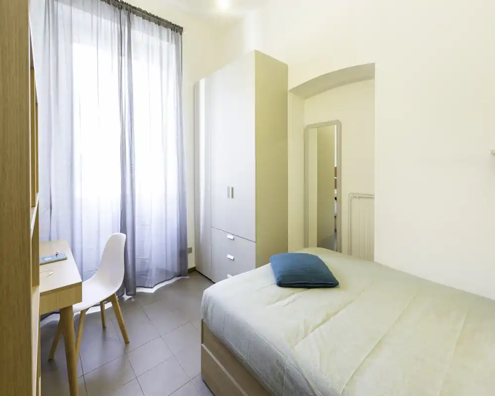 Camera da letto luminosa e minimale con letto ben fatto, armadio e una scrivania vicino alla finestra — ideale per un soggiorno confortevole per una o due persone.