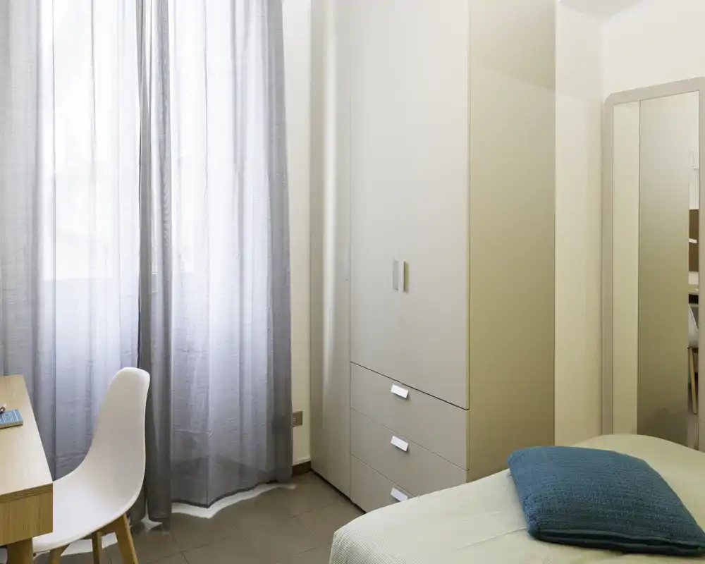 Angolo camera luminoso e ordinato con armadio, scrivania e letto parzialmente visibile con cuscino turchese — stile moderno e tranquillo, ideale per soggiorni brevi.