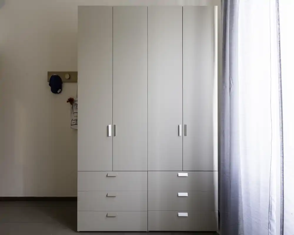 A minimal, well-lit shot focused on a white wardrobe/closet with drawers; sheer curtains are visible at the right edge and a small coat hook with a hat on the left wall.