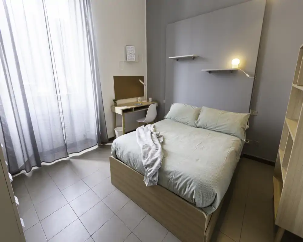 Bright, modern bedroom with a double bed, neat linens, bedside lamp and a small study desk by a large window with sheer curtains.