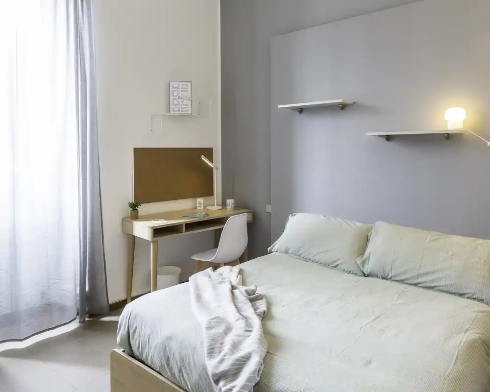 Well-lit, modern bedroom with a double bed, soft linens and a small wooden desk by the window — calm, neutral tones and simple shelving create a serene space.