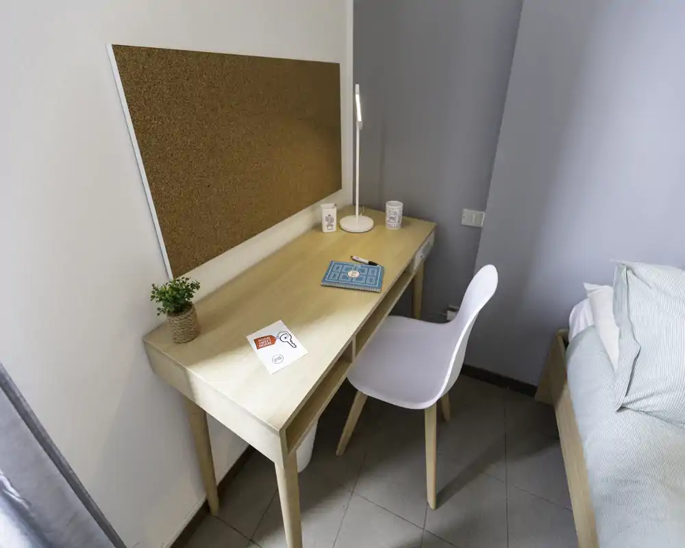 Bright, minimal bedroom corner with a light wood desk, cork notice board and a white chair beside a neatly made bed. The space shows a tidy study spot integrated into the sleeping area with soft, natural lighting.