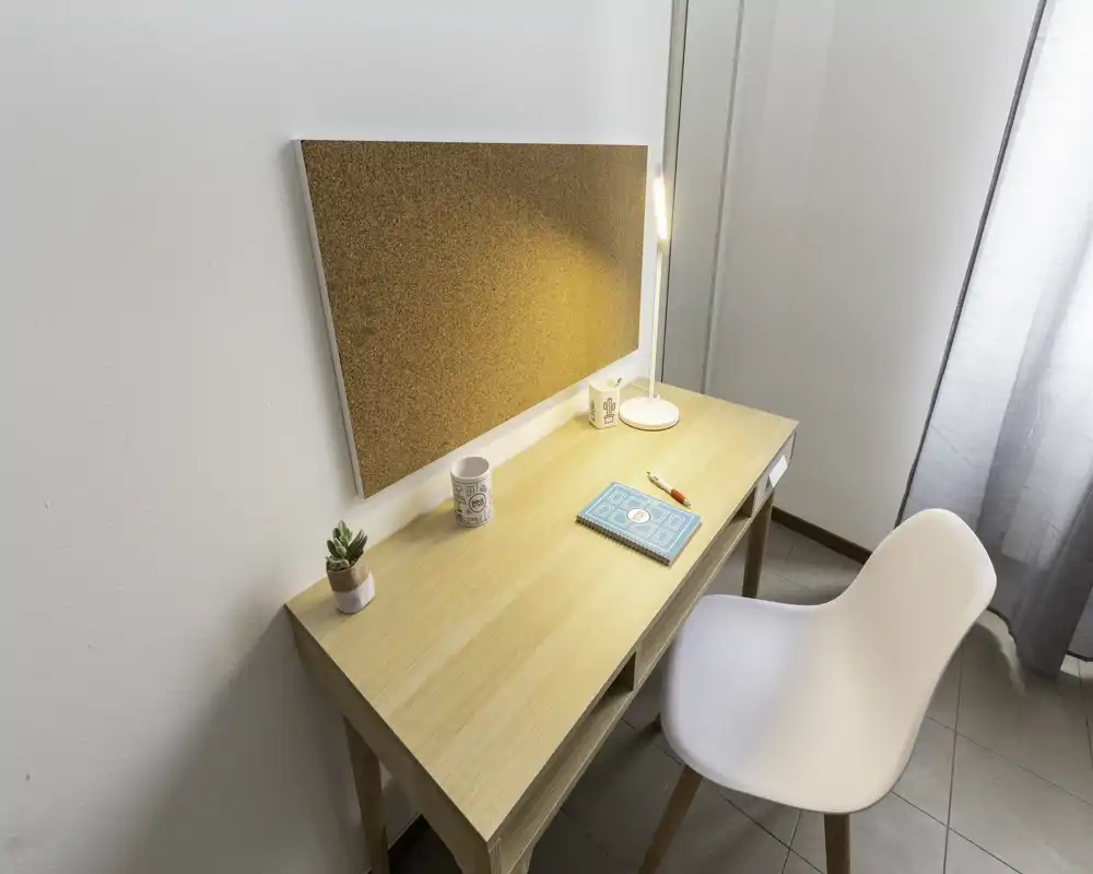 Angolo scrivania luminoso e minimale con bacheca in sughero, lampada, piantina e sedia bianca — perfetto come piccolo spazio studio o home office.