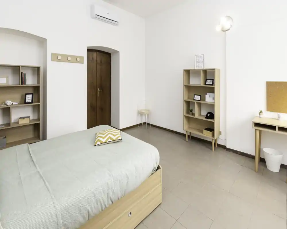 Camera da letto luminosa e minimal con un letto matrimoniale ben rifatto, mobili in legno chiaro e biancheria verde tenue. Ambiente arioso e ordinato con scaffali e una piccola scrivania.