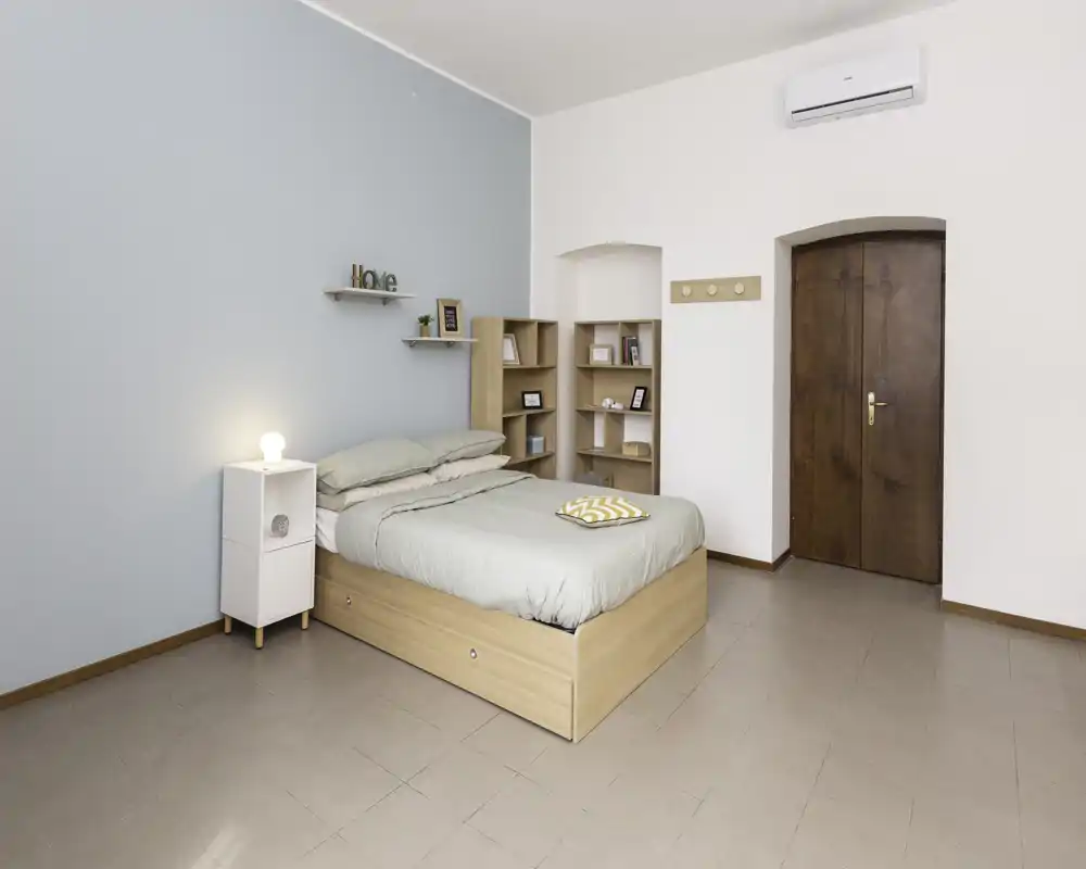 Camera da letto luminosa e minimal con letto in legno completo, biancheria verde chiaro, comodino con lampada e mensole semplici; parete pastello e porta in legno creano un'atmosfera tranquilla perfetta per gli annunci.