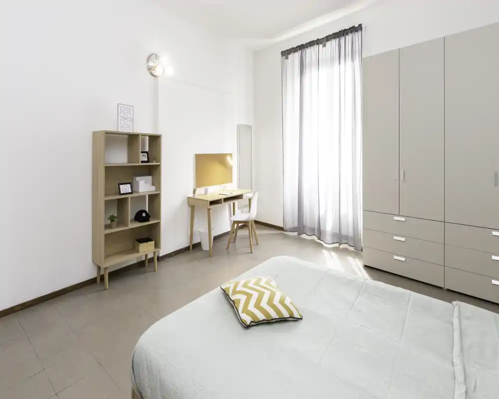 Camera da letto luminosa e minimal con letto ben rifatto, armadio, scaffale e una scrivania vicino a una finestra che lascia entrare luce naturale.