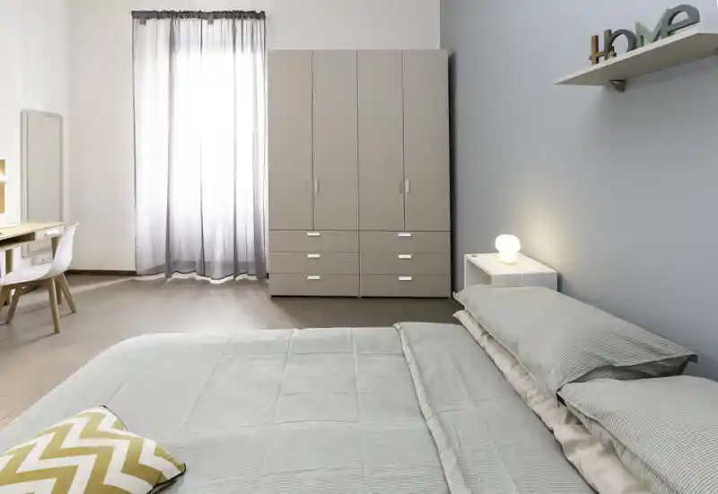 Camera da letto moderna e rilassante con letto matrimoniale ben rifatto, biancheria verde tenue, comodino con lampada e un grande armadio vicino alla finestra.